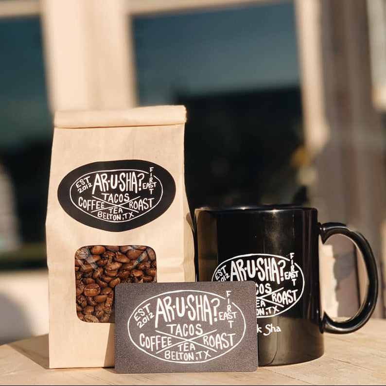 Arusha Coffee Tea Roast coffee bag, black mug, and business card on a wooden surface with a blurred outdoor background.